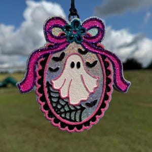 May include: A hanging oval ornament featuring a white ghost, black spiderweb, and small black bats on a silver glitter background. The frame is outlined in bright pink glitter with a pink glitter bow and a small teal flower at the top center.