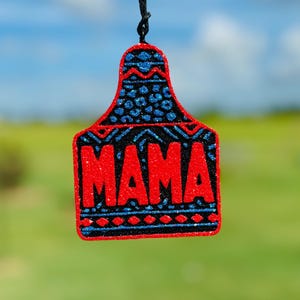 May include: A red and blue glittery ornament with the word "MAMA" in red letters. The ornament is shaped like a cow tag and has a black string attached to the top.