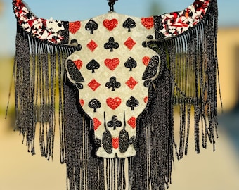 Peut inclure: Un crâne de taureau blanc avec des franges noires et un motif de cartes à jouer rouge et noir. Le crâne est suspendu à un cordon noir avec une boule de strass rouge.