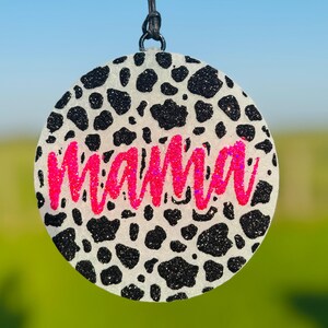 May include: Round ornament with a cow print design in black and white. The word "mama" is written in bright pink glitter across the center. The ornament hangs from a black string.