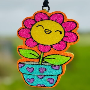 May include: A cheerful, pink and yellow flower with a smiling face is potted in a blue and purple pot with hearts. The flower is made of glitter foam and is hanging from a black string.