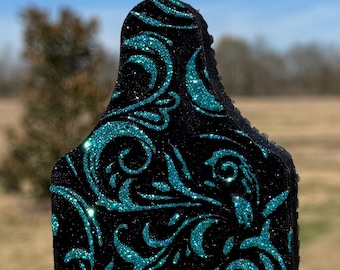 May include: A black livestock ear tag ornament featuring a vibrant teal glitter filigree pattern. The tag is shaped with a rounded top and a rectangular base, suspended by a thin black cord attached to a small metal ring at the apex.