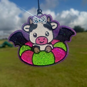 May include: A glittery hanging ornament featuring a cartoon cow with black and white markings, wearing a white bow, and sporting black bat wings. The cow is positioned inside a bright pink and lime green glittery inflatable pool float.