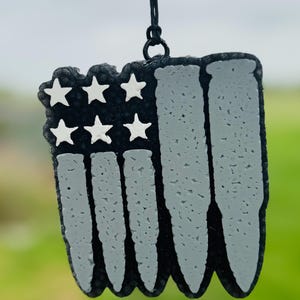 May include: A black and gray American flag ornament with a string for hanging. The flag is made of bullet casings and has white stars.