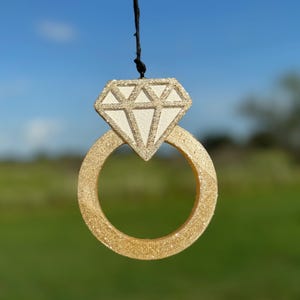 May include: A gold ring with a sparkly diamond on top. The ring is hanging from a black string against a blue sky background.