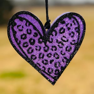 May include: A purple heart-shaped ornament with a black glitter outline and leopard print design. The ornament is suspended by a black string. The background is blurred, suggesting an outdoor setting.