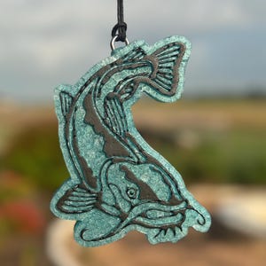 May include: A hanging ornament shaped like a catfish, featuring a textured, translucent teal body with black detailing along the fins, tail, and head. The fish is suspended by a thin black cord attached to a silver-toned metal jump ring at the top.