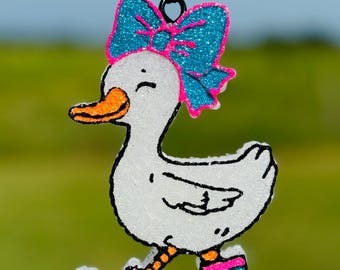 May include: A white duck ornament with a teal glitter bow and matching boots. The duck has an orange beak and legs, and is outlined in black. The boots have a blue leopard print with pink trim. The ornament hangs from a black string.