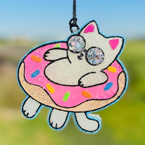 May include: A white cat wearing sunglasses floats in a pink donut-shaped pool float with colorful sprinkles. The cat has a white body with black eyes and a pink nose. The donut float has a pink center with blue sprinkles and a light blue outer ring.
