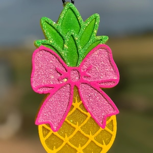 May include: A glittery pineapple-shaped ornament with a pink bow. The pineapple is yellow with a textured pattern, and the leaves are green. The bow and leaves have glitter accents. A black string is attached for hanging.