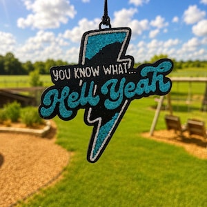 May include: A lightning bolt-shaped hanging ornament with a black base and teal glitter accents. The text "YOU KNOW WHAT... Hell Yeah" is printed in a silver glitter font across the center of the bolt. It hangs from a black cord.