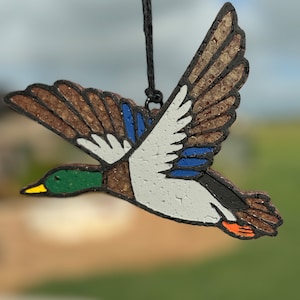 May include: A brown, white, and blue painted wooden duck ornament hanging from a black string. The duck is flying with its wings spread out. The duck has a green head and a yellow beak.