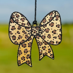 May include: A decorative bow ornament featuring a gold and black leopard print pattern with a glittery texture. The bow is outlined in black and hangs from a thin black string attached to a small metal loop at the center of the bow structure.