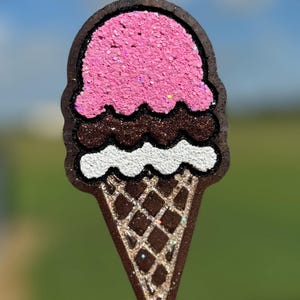 May include: A glittery ice cream cone-shaped pendant. The top scoop is pink, the middle is chocolate, and the bottom is a waffle cone. The pendant is brown with glitter accents and a black cord.