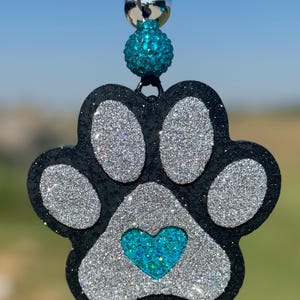 May include: A hanging ornament shaped like a paw print with a black base, silver glitter pads, and a central turquoise glitter heart. The top features a silver metallic bead and a textured turquoise bead attached to a black cord for hanging.