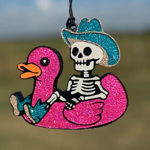 May include: A glittery hanging ornament featuring a white skeleton wearing a teal cowboy hat and matching boots, sitting inside a bright pink glittery rubber duck floatie. The ornament is suspended by a thin black string against a blurred background.