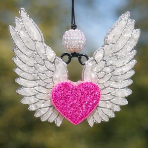 May include: A hanging ornament featuring a bright pink glitter heart centered between two white glittery angel wings. Above the heart, a small iridescent sphere is attached to a black cord, creating a decorative piece with a sparkling finish.