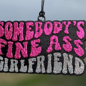 May include: Black rectangular ornament with the text "SOMEBODY'S FINE ASS GIRLFRIEND" in pink and silver glitter lettering. The ornament is hanging from a black string.