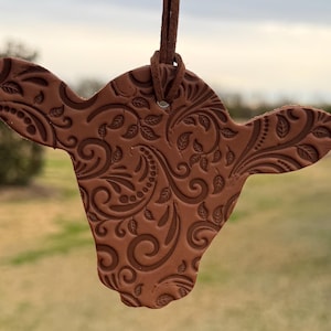 May include: A brown leather cow head ornament with a paisley design. The ornament is hanging from a brown cord.