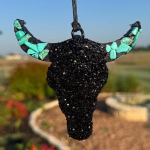 May include: Black glitter bull skull pendant with turquoise accents on the horns. The pendant is suspended from a black cord. The background is blurred, showing a natural outdoor setting.
