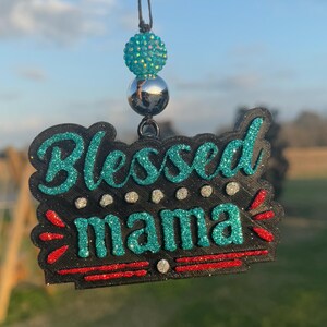 May include: A black car charm with the words "Blessed mama" in teal glitter. The charm has a red and white accent and a teal and silver beaded top.