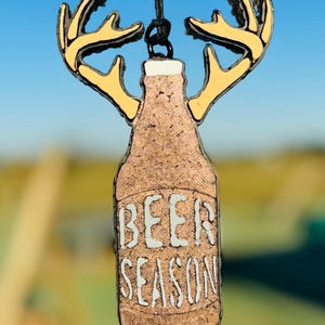 May include: A wooden ornament shaped like a beer bottle with antlers on top. The bottle is brown and has the words "BEER SEASON" written on it in white.