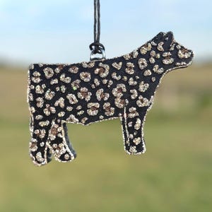 May include: A black cow-shaped ornament with a leopard print pattern. The ornament is outlined with glitter and suspended by a black string. The background is a blurred view of a field.