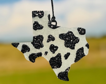 May include: A Texas-shaped ornament with a cow print design. The ornament is white with black glitter spots and a black string for hanging. The background is blurred with blue and green hues.