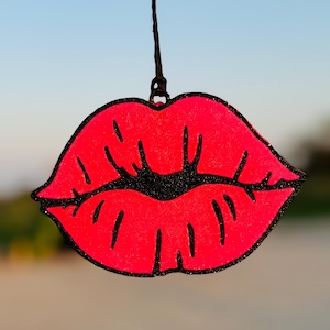 May include: A bright pink glitter lips ornament with a black outline. The ornament is hanging from a black string.