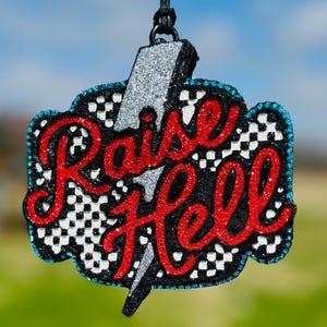 May include: A hanging ornament featuring the text Raise Hell in red glitter script. The background is a black and white checkered pattern with a silver glitter lightning bolt overlay. The entire piece is outlined in small, bright blue rhinestones.