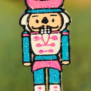 May include: A glittery, colorful nutcracker ornament. The ornament features a blue and pink hat, blue jacket, and pink accents. The face is tan with a white mustache. The ornament is strung on a black cord, perfect for holiday decor.