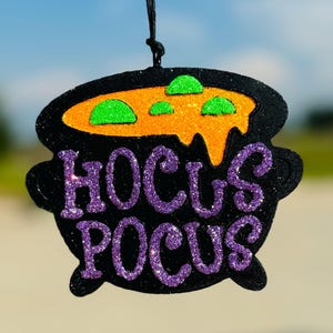 May include: Black cauldron-shaped decoration with orange and green glitter accents. The cauldron has the words "Hocus Pocus" in purple glitter. A black string is attached for hanging. The background is blurred.