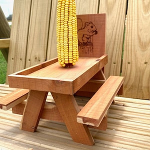 May include: A miniature wooden picnic table with a corn cob on top and a squirrel design on the back. The table is made of natural wood and has a rustic look.