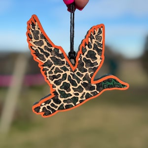 May include: A wooden duck ornament with an orange outline, black and tan patterned wings, and a green head. The ornament is suspended by a black string. The background is blurred, suggesting an outdoor setting.
