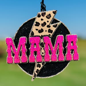 May include: A circular hanging ornament featuring a black glitter background with a central beige lightning bolt patterned with brown leopard spots. The word MAMA is written in large, bright neon pink glitter letters across the center.