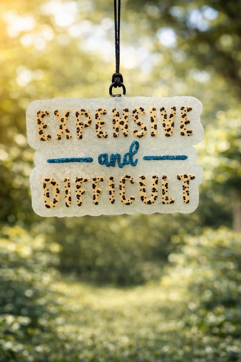 May include: A glittery, rectangular sign with the words "EXPENSIVE" and "DIFFICULT" in a leopard print font. The word "and" is written in cursive with a blue glitter underline. The sign hangs from a black cord.