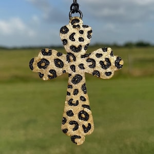 May include: A gold-colored cross pendant with a black leopard print pattern. The cross is suspended from a black cord. The background is a blurred view of green grass and a blue sky.