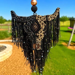 May include: A hanging ornament shaped like a bull skull, featuring a split design with black stones on one side and a gold leopard print on the other. It is adorned with long black fringe, small iridescent beads, and topped with a large peach bead.