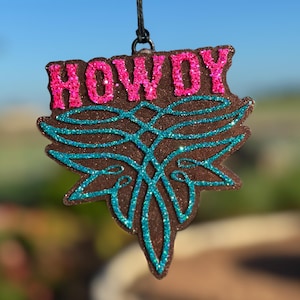 May include: A brown, glittery ornament with the word "HOWDY" in pink glitter. The ornament has a turquoise glitter design and is suspended by a black string. The ornament has a decorative shape.