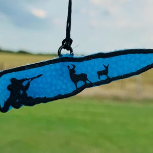 May include: A bullet-shaped hanging ornament featuring a bright blue translucent center with a black outline. The design depicts a silhouette of a person aiming a rifle at two deer standing on a textured surface, suspended by a black cord.
