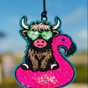 May include: A brown and black Highland cow wearing green sunglasses and riding a pink flamingo pool float. The cow and flamingo are made of glitter and have a blue outline.