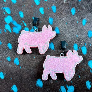 May include: Two pink glitter pig-shaped earrings with silver outlines and black clip-on closures. The earrings are set against a dark background with blue splatter paint.