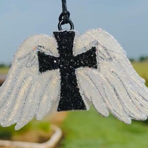 May include: A decorative ornament featuring angel wings and a cross. The wings are white with silver glitter, and the cross is black with glitter. The ornament is suspended by a black cord.