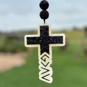 May include: A black glitter-filled cross pendant with a white outline and the text "<GAV>". The cross is suspended from a black beaded string with a black sphere and a textured bead. The background is blurred green.
