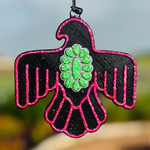 May include: Black pendant in the shape of a bird, with pink glitter accents and a green and silver center design. The bird has outstretched wings and is suspended by a black cord. The pendant is likely a decorative item or accessory.