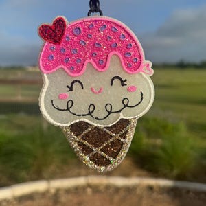 May include: A hanging ornament shaped like a smiling ice cream cone with a glittery pink scoop topped with a red heart, a white middle section featuring a stitched face, and a brown waffle cone base with gold glitter accents and a black loop.