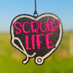 May include: Black heart-shaped ornament with a white outline and a stethoscope design. The words "SCRUB LIFE" are written in pink glitter.