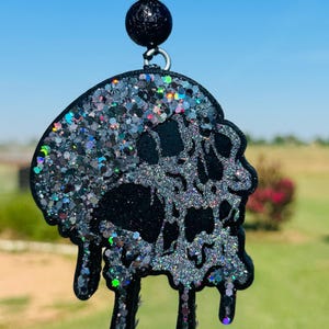 May include: A black skull-shaped car charm with a glittery, iridescent finish. The charm has a dripping design and is suspended from a black beaded chain. The background is a blurred view of a sunny day.