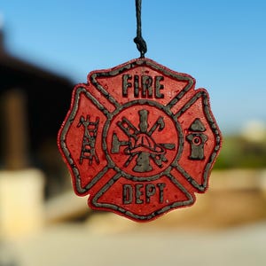 May include: A red and black hanging ornament shaped like a Maltese cross, featuring the text FIRE DEPT. The center displays a fire helmet, ladder, and hydrant icon, all rendered in a textured, raised relief design suspended by a black string.