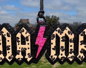 May include: A hanging ornament featuring the word MAMA in a bold, black-outlined font. The letters have a beige base with a brown leopard print pattern and glitter accents. A bright pink glitter lightning bolt replaces the letter A.